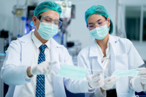 Two factory worker man and woman responsible to check quality control of product in front of machine of mask industry.